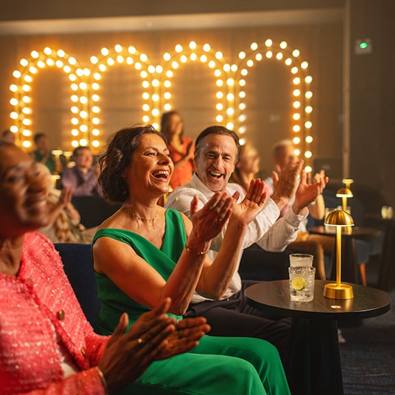 Angie clapping excitedly in the foreground at the entertainment as Mick stares at her lovingly in the background